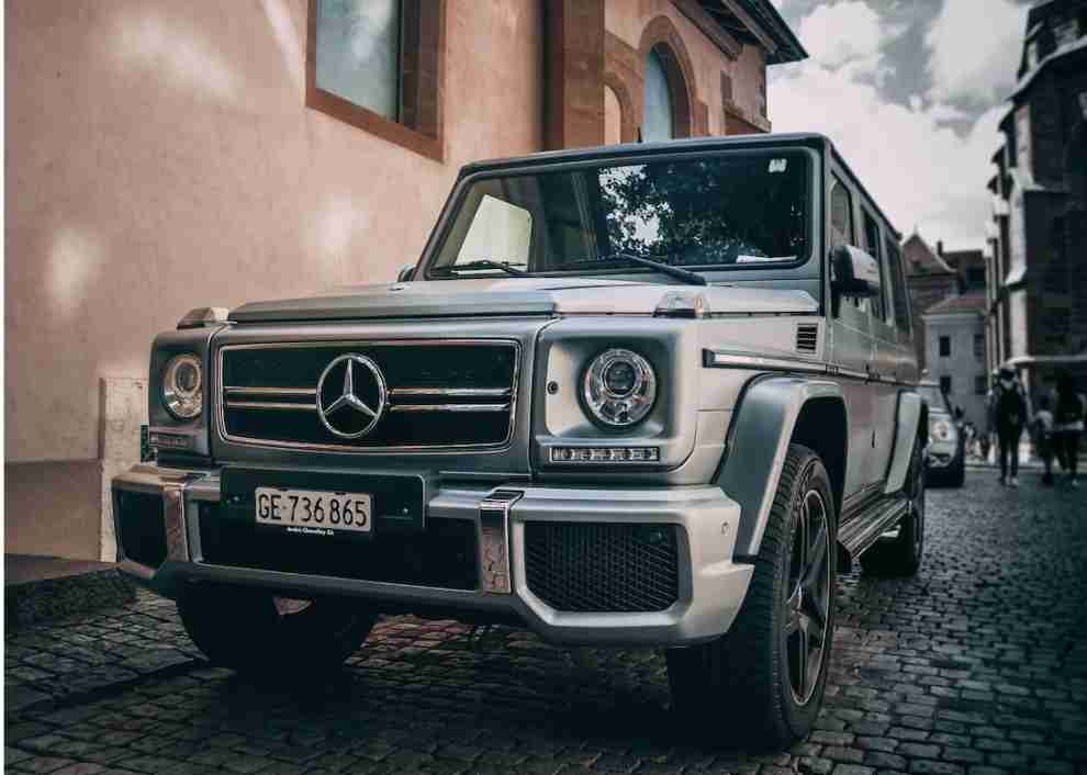 A silver Mercedes-Benz G-Class SUV parked on a cobblestone street near historical buildings.